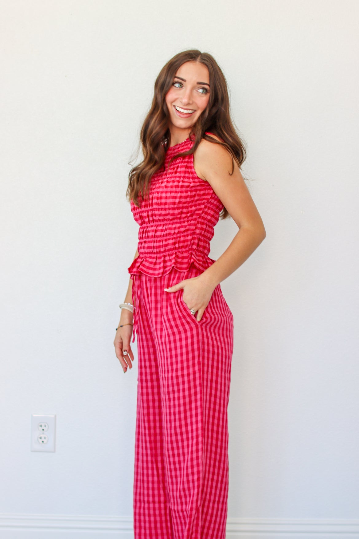 A woman modeling a sleeveless red and pink gingham tank and pant set with a smocked waist and ruffle detail, standing against a plain white wall.