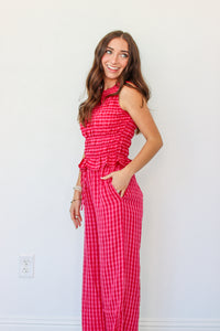 A woman modeling a sleeveless red and pink gingham tank and pant set with a smocked waist and ruffle detail, standing against a plain white wall.