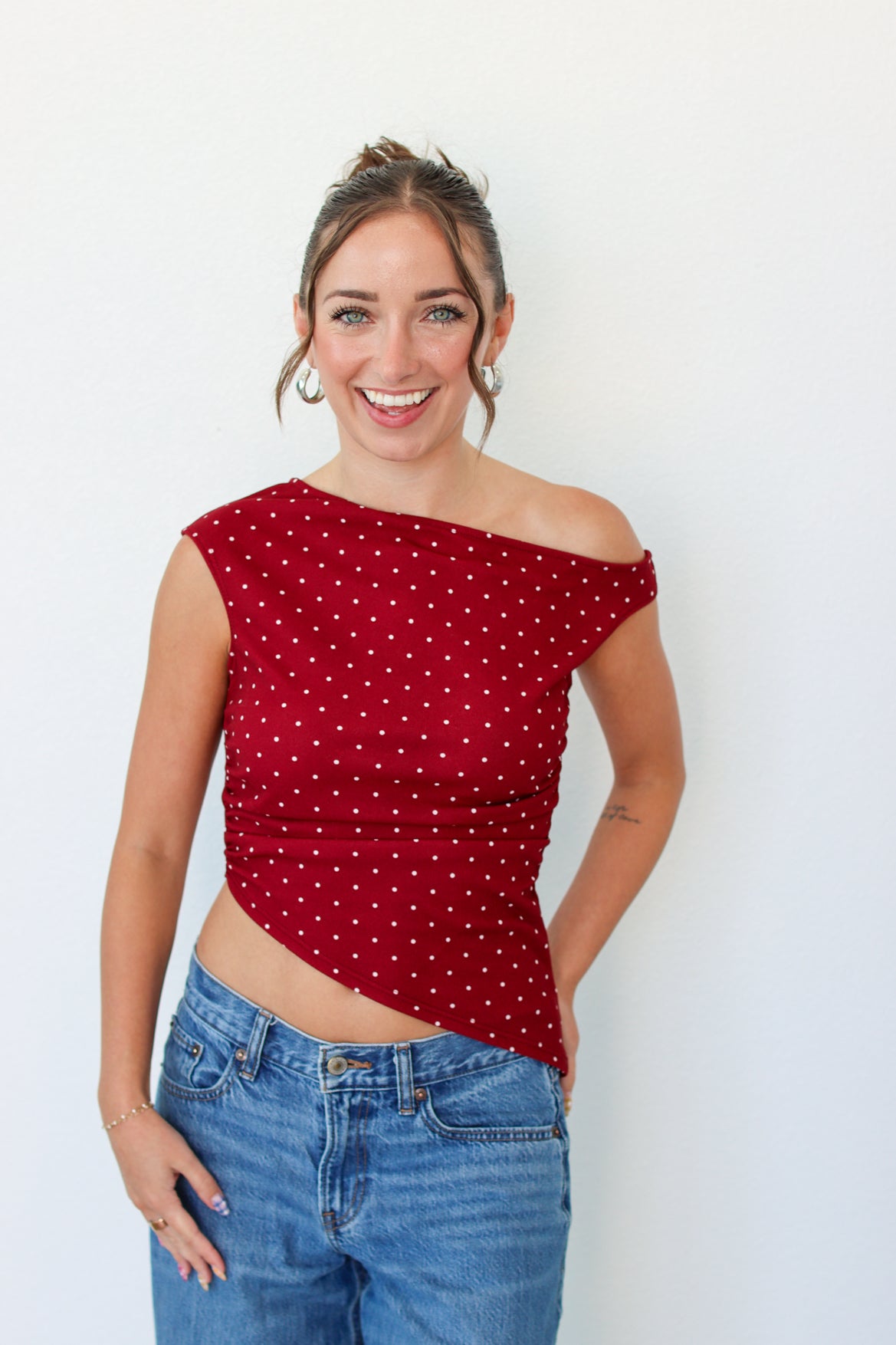 A woman smiling and wearing a red off-the-shoulder crop top with white polka dots, paired with blue jeans, standing against a plain white wall.