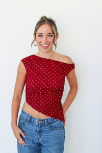 A woman smiling and wearing a red off-the-shoulder crop top with white polka dots, paired with blue jeans, standing against a plain white wall.