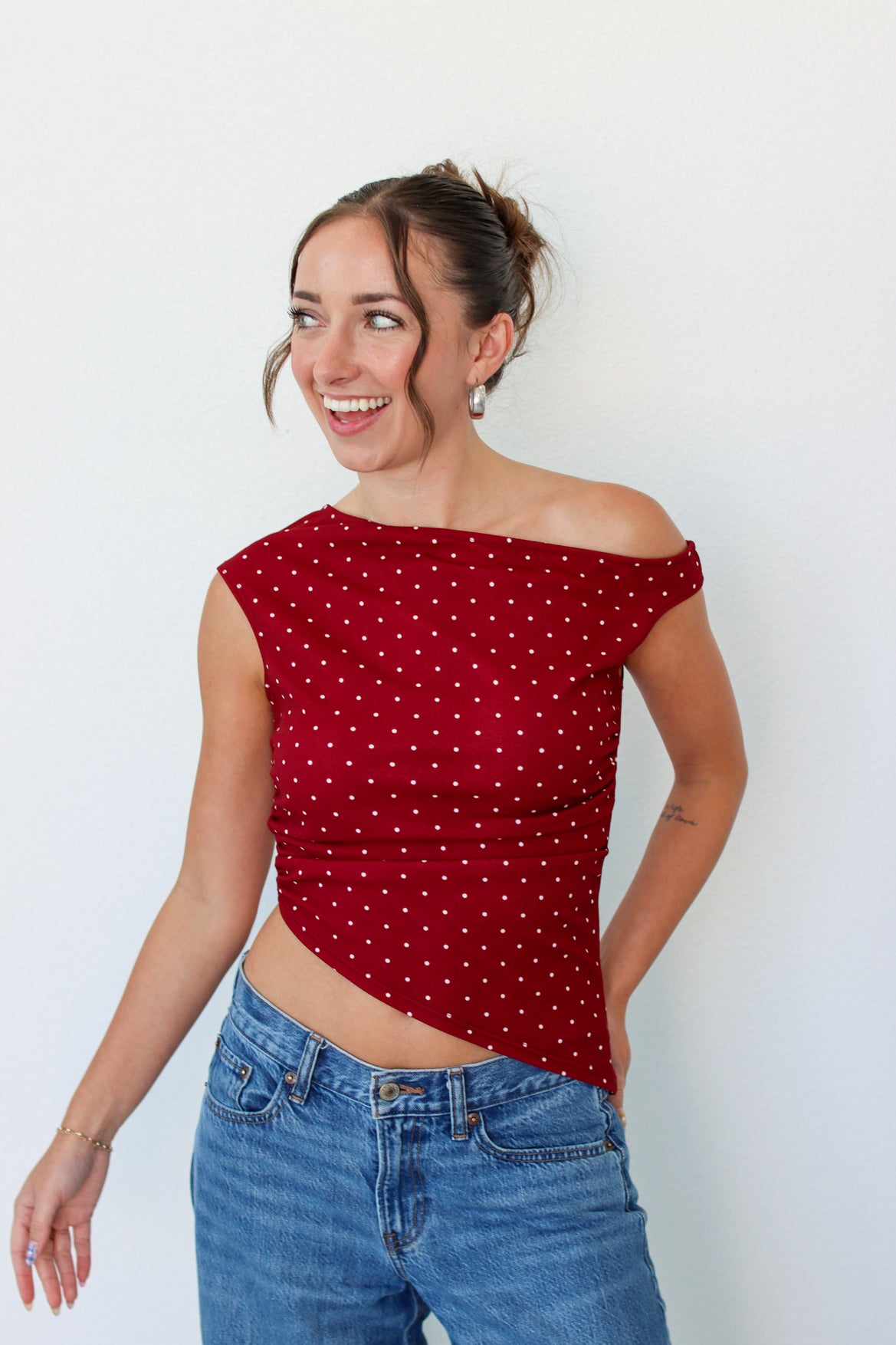 A woman wearing a red off-the-shoulder crop top with white polka dots and blue jeans, standing against a plain white wall.