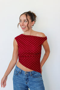 A woman wearing a red off-the-shoulder crop top with white polka dots and blue jeans, standing against a plain white wall.