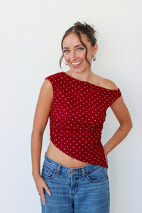 A woman wearing a red off-the-shoulder crop top with white polka dots and blue jeans, standing against a plain white wall.
