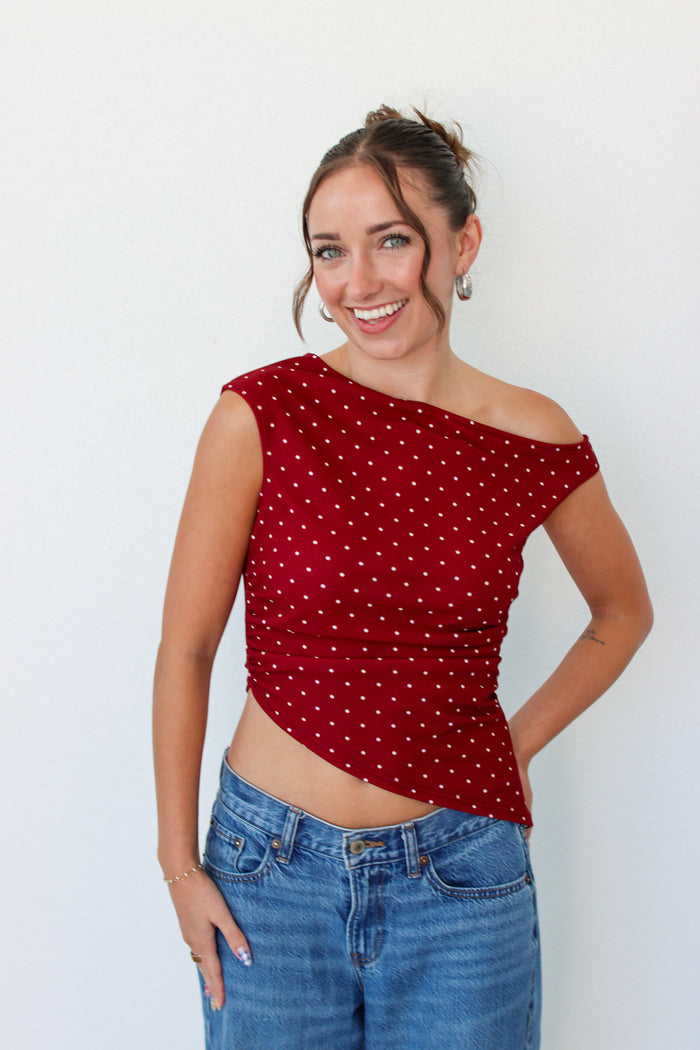 A woman wearing a red off-the-shoulder crop top with white polka dots and blue jeans, standing against a plain white wall.