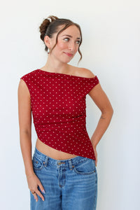 A woman modeling a red off-the-shoulder top with small white polka dots, paired with blue jeans, against a plain white background.