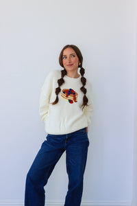 Woman wearing a white sweater with a colorful design and blue jeans against a white background