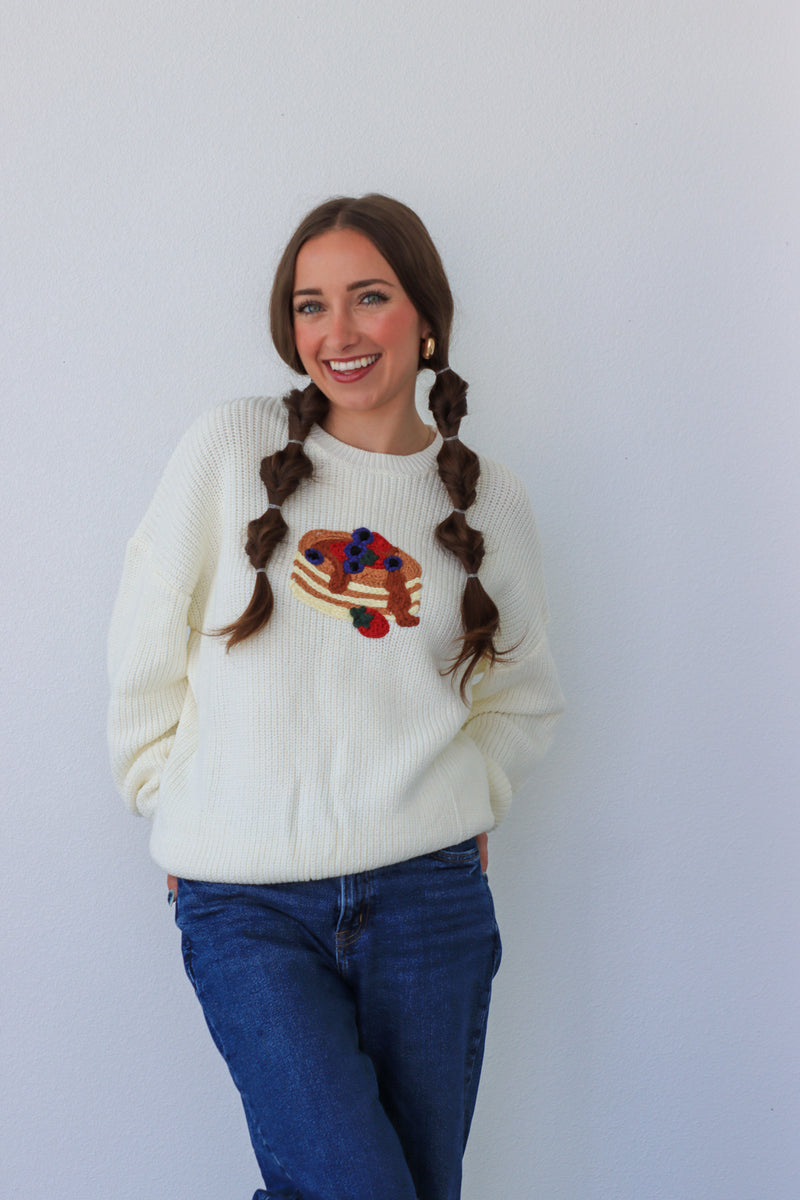 Woman wearing a cream sweater with a colorful design, standing against a plain background