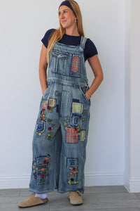 Woman wearing a denim jumpsuit with colorful patches against a white wall.