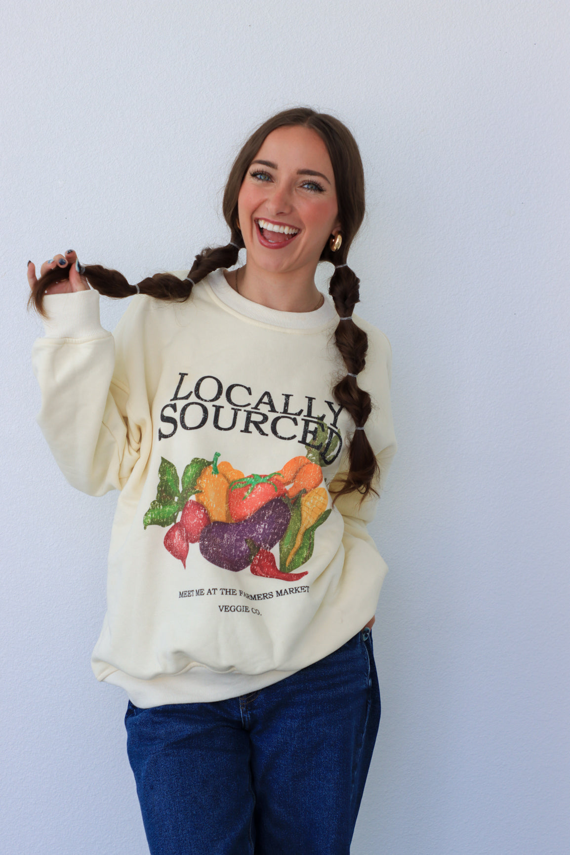 Woman wearing a cream-colored sweatshirt with 'Locally Sourced' graphic, standing against a light blue background.