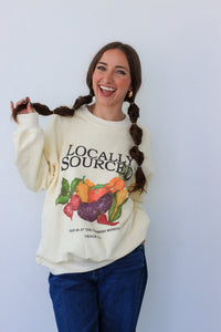 Woman wearing a cream-colored sweatshirt with 'Locally Sourced' graphic, standing against a light blue background.