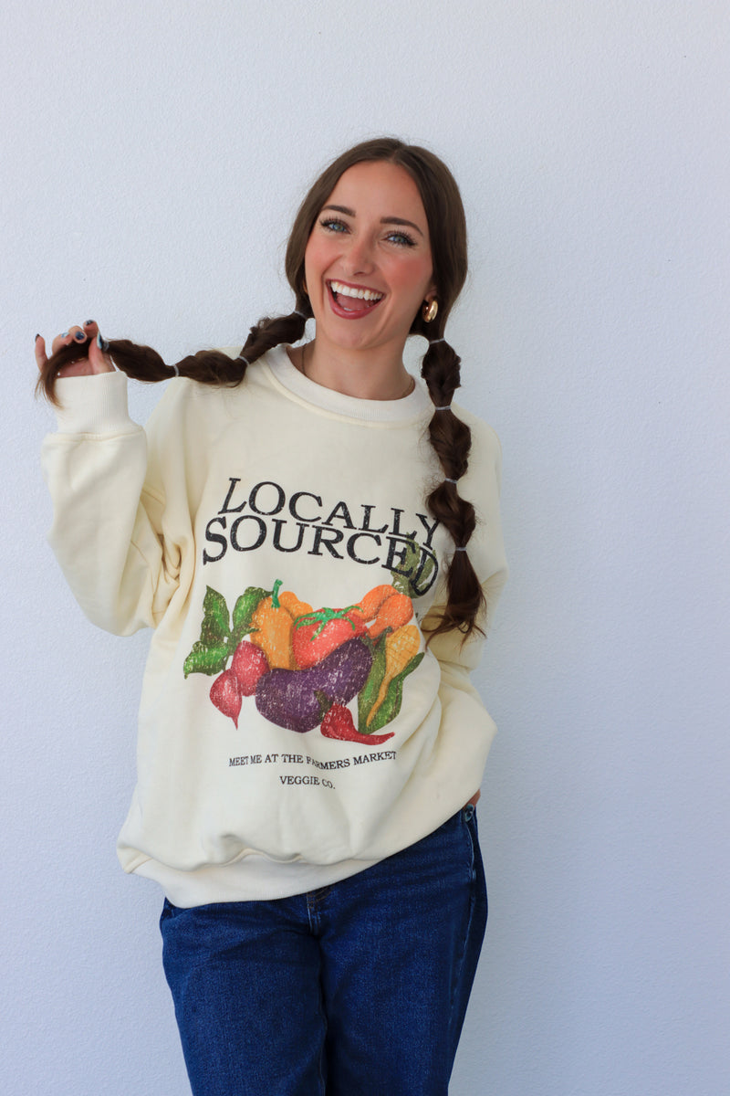 Woman wearing a cream-colored sweatshirt with 'Locally Sourced' graphic, standing against a light blue background.