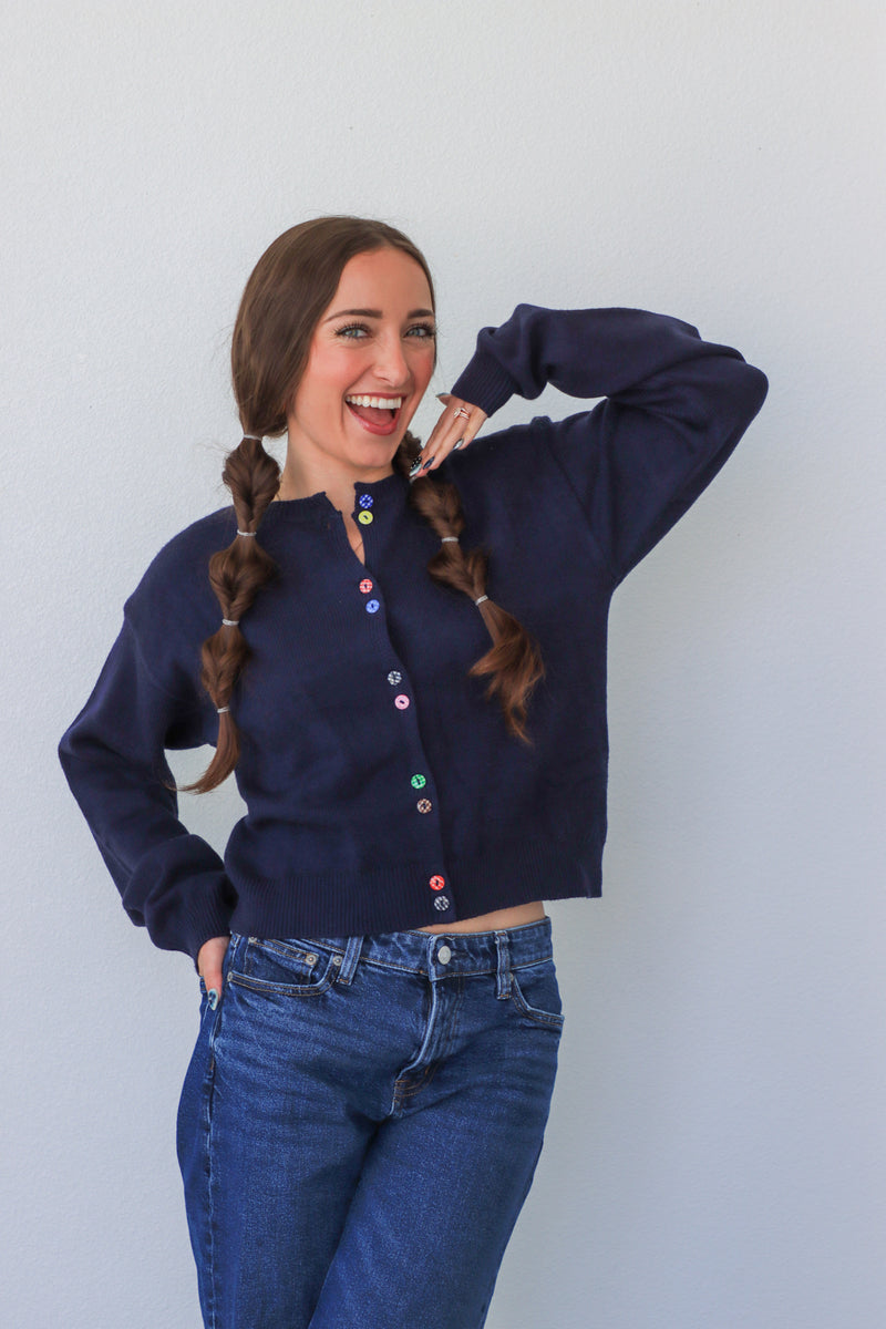 Woman wearing a navy cardigan with colorful buttons and blue jeans against a white background