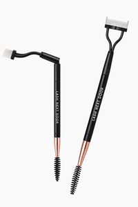 Two black lash tools with rose gold ferrules and white bristles, shown on a light gray background. One is labeled 'LASH NEXT DOOR' and has a curved arm with a small brush attachment, and the other is a spoolie-style lash tool with a black T-handle.