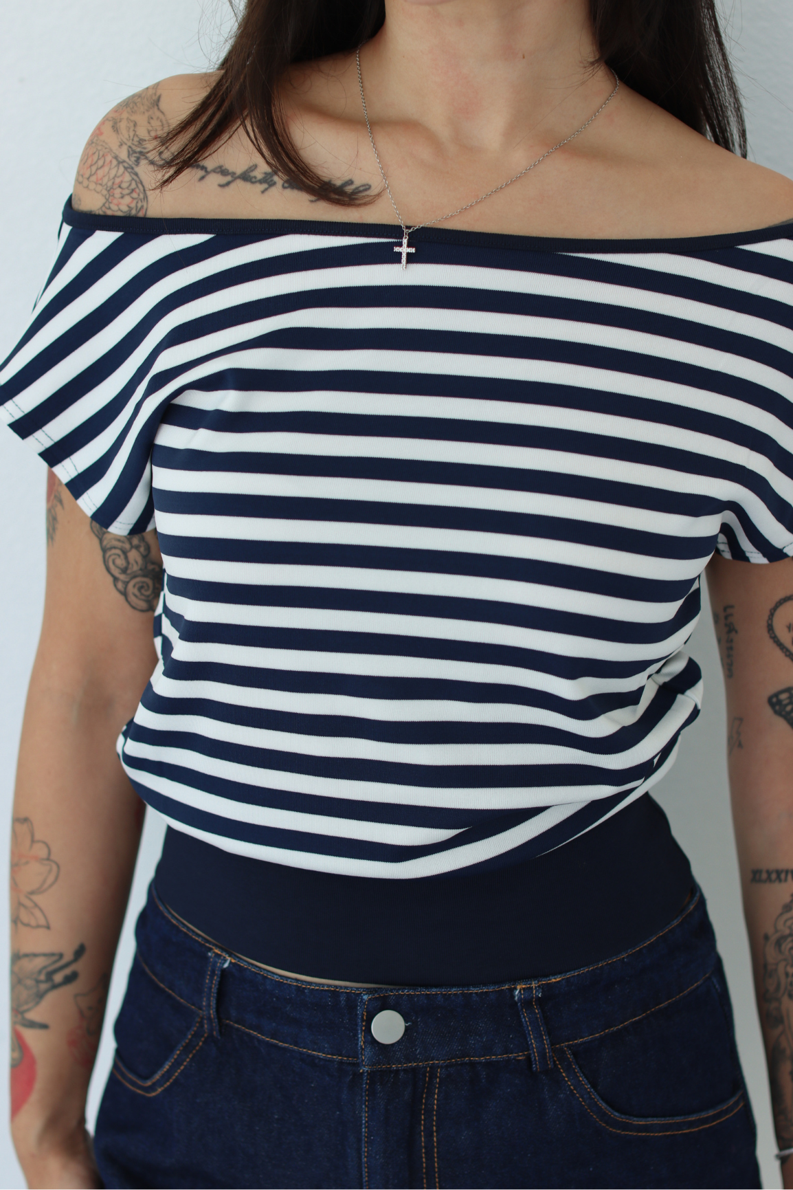 A woman wearing a navy and white horizontally striped off-the-shoulder top with a wide navy waistband, paired with dark blue jeans.