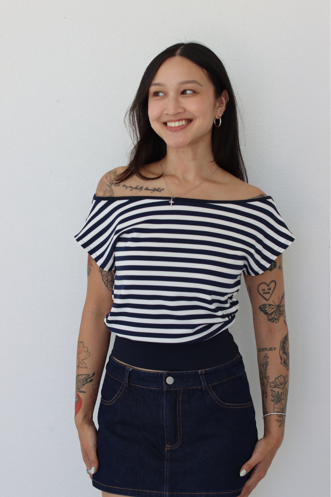 A woman with tattoos wearing a navy and white horizontally striped off-the-shoulder t-shirt and a dark blue denim mini skirt, standing against a plain white wall.