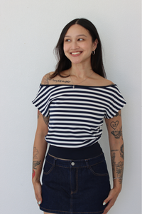 A woman with tattoos wearing a navy and white horizontally striped off-the-shoulder t-shirt and a dark blue denim mini skirt, standing against a plain white wall.