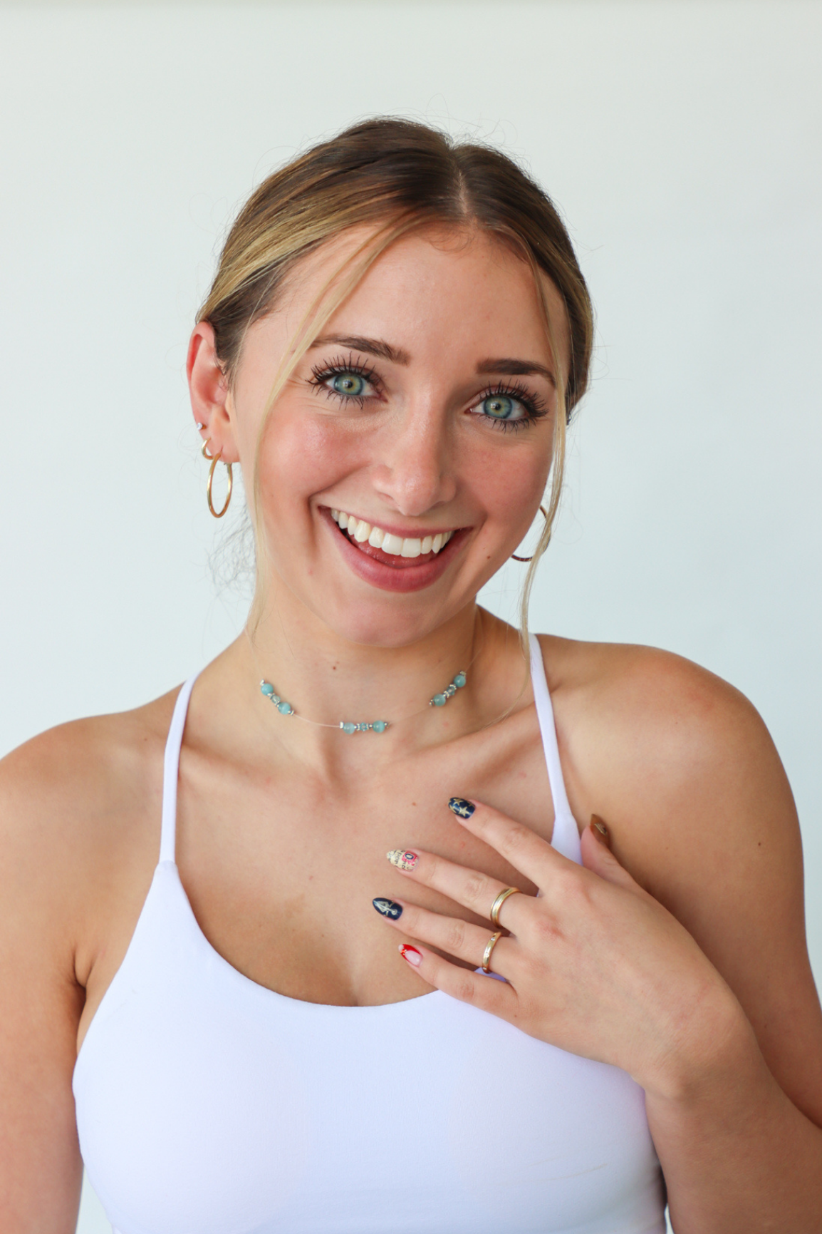 Woman wearing a white tank top and jewelry, smiling against a plain background