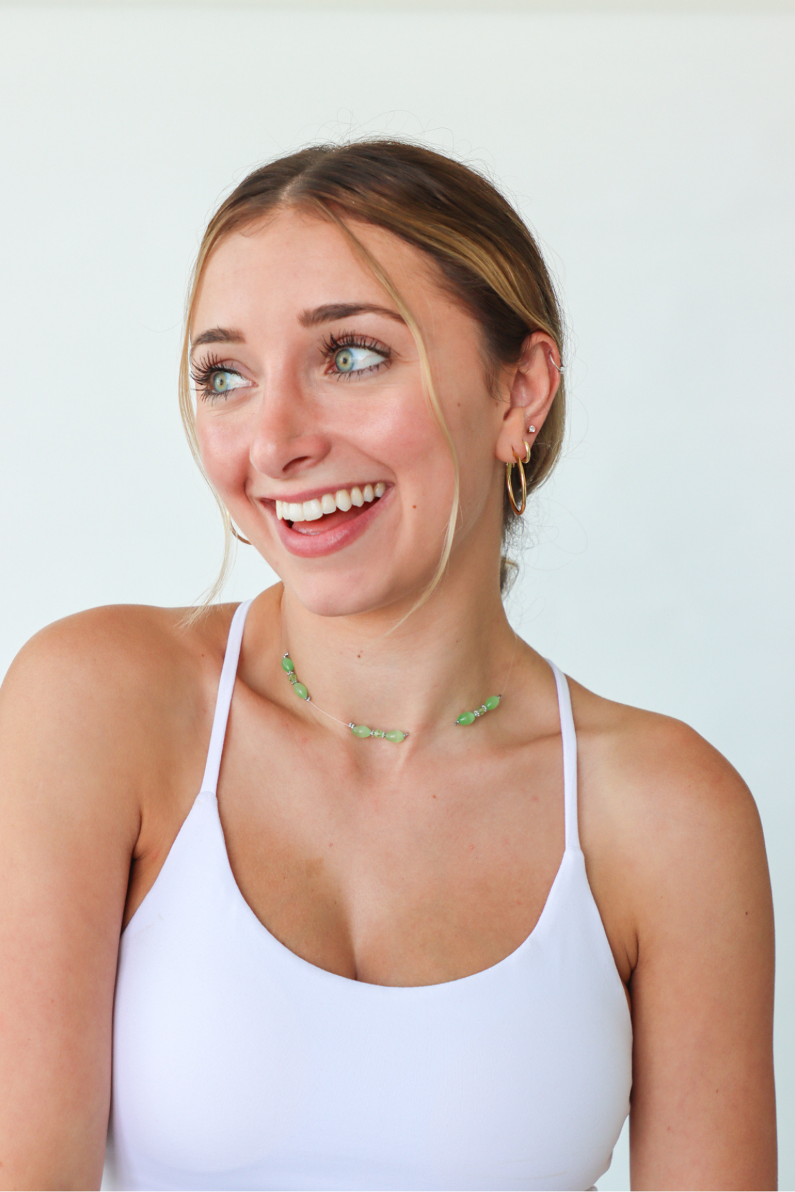 Woman wearing a white tank top and green necklace against a plain background