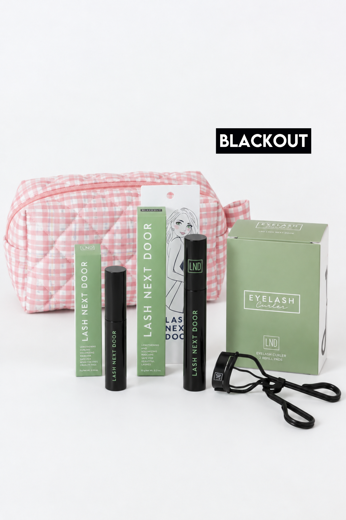 Lash Next Door eyelash extension kit with a pink and white gingham cosmetic bag, black mascara tubes, a green product box, and a black strap, displayed on a white background with a 'BLACKOUT' label.