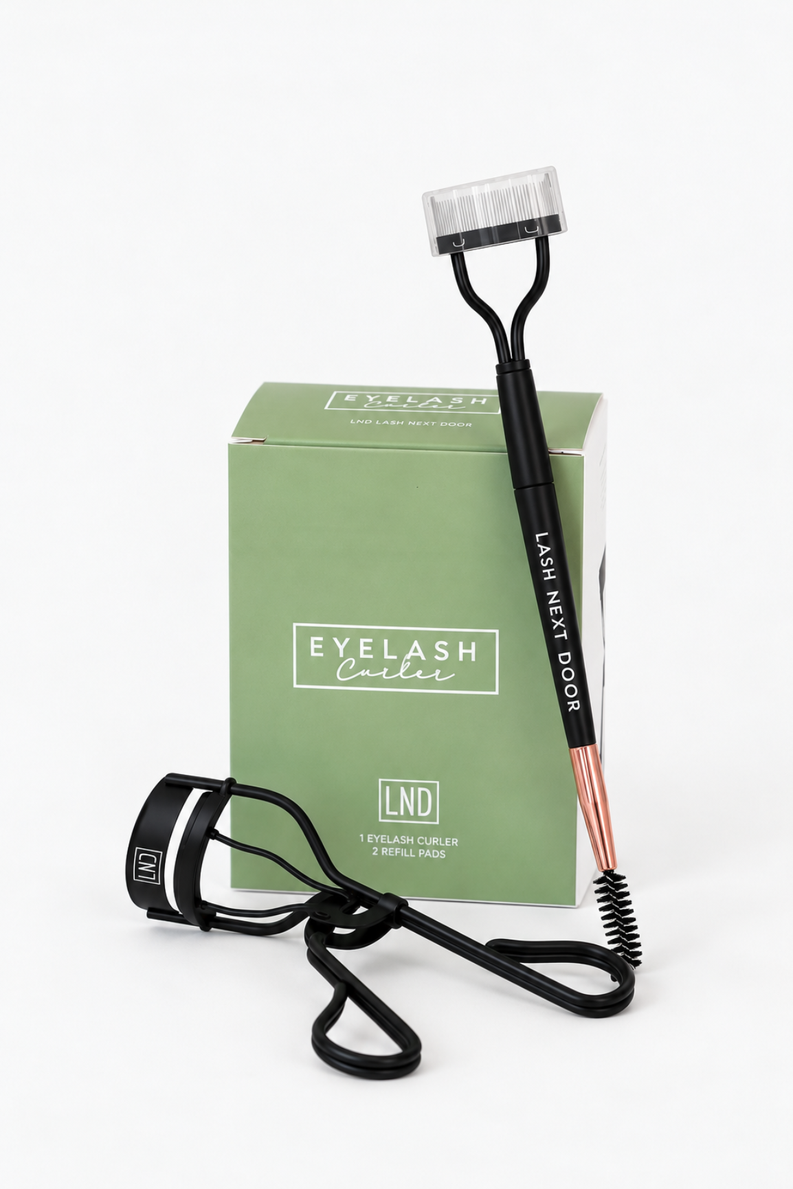An eyelash curler set with a black eyelash curler, a black spoolie-style brow tool with rose gold ferrule, and a sage green product box labeled 'Eyelash Curler' and 'LND'.
