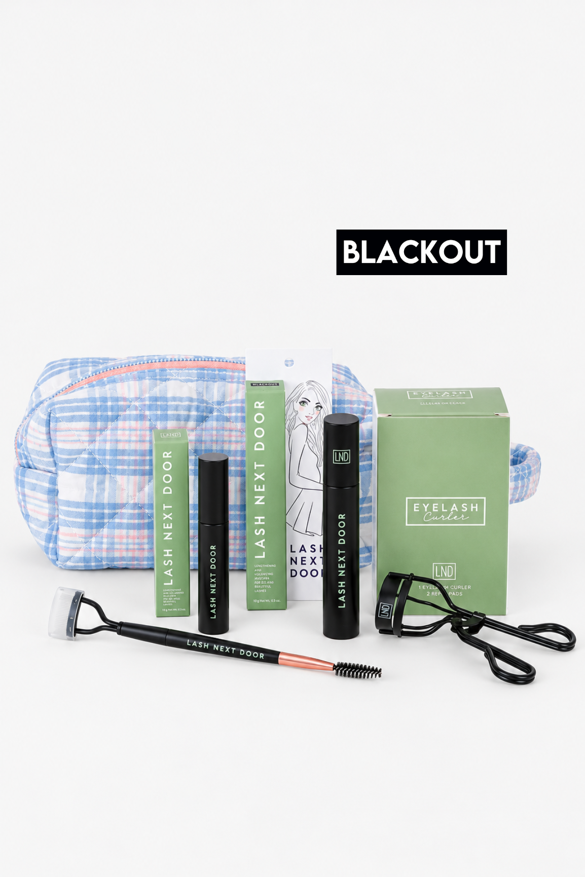 Eyelash extension kit with black lash adhesive, black lash primer, a green lash box, a green instruction booklet, a blue and white plaid makeup bag, a black spoolie, and a black lash comb, arranged on a white background with a black 'BLACKOUT' label above.
