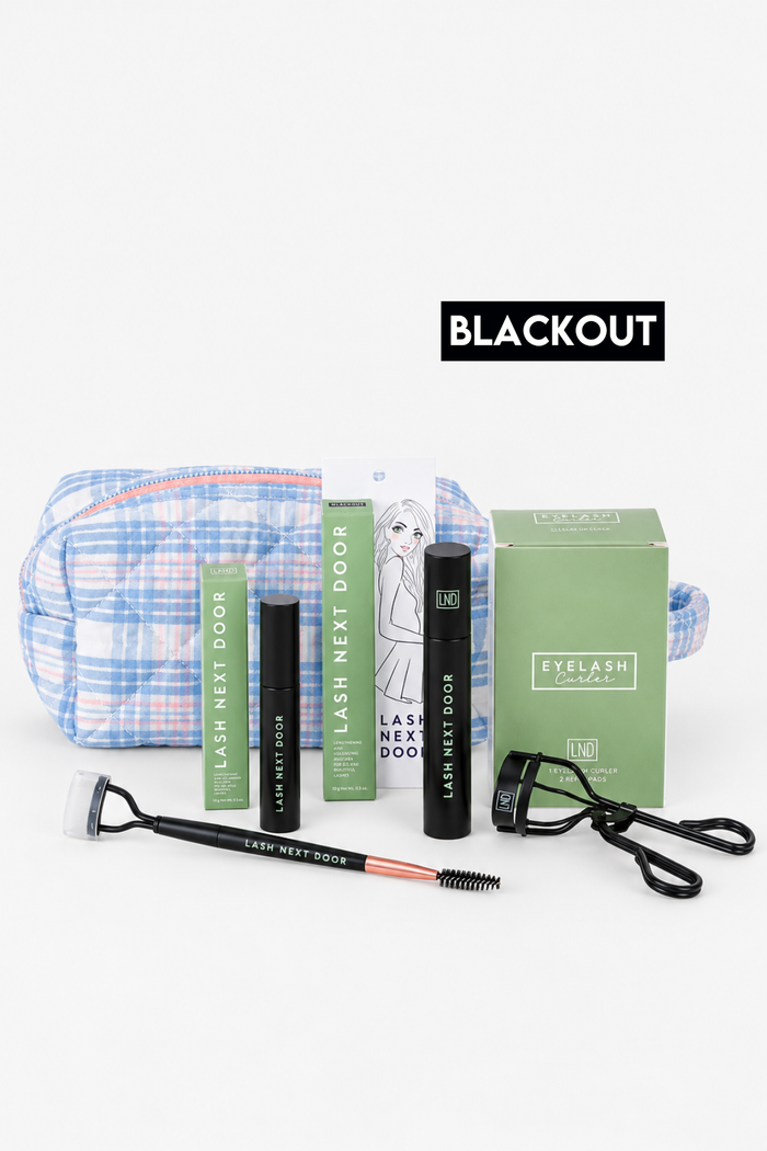 Eyelash extension kit with black lash adhesive, black lash primer, a green lash box, a green instruction booklet, a blue and white plaid makeup bag, a black spoolie, and a black lash comb, arranged on a white background with a black 'BLACKOUT' label above.