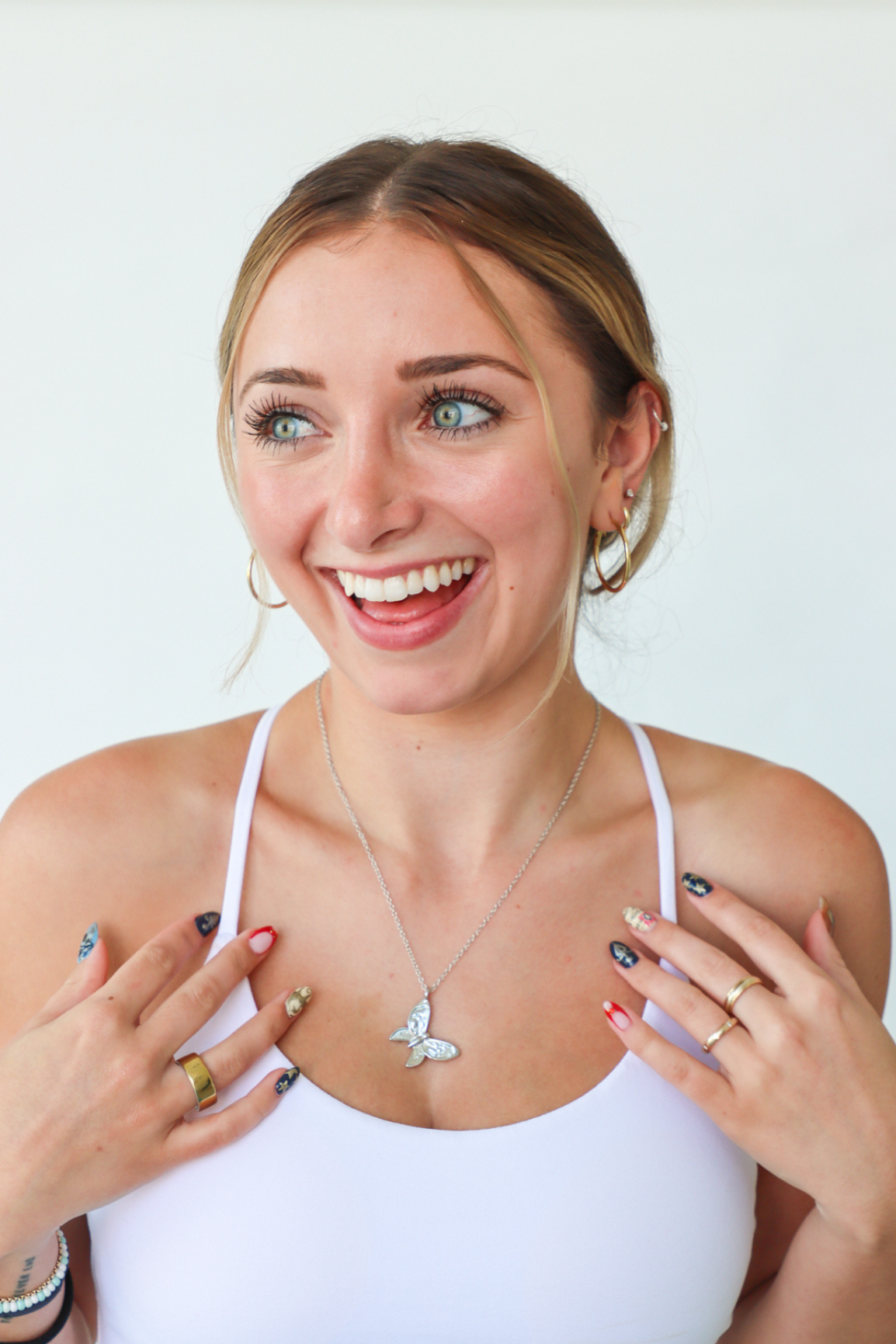 Woman wearing a white top with jewelry, including a necklace and rings, on a plain background.