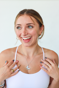Woman wearing a white top with jewelry, including a necklace and rings, on a plain background.