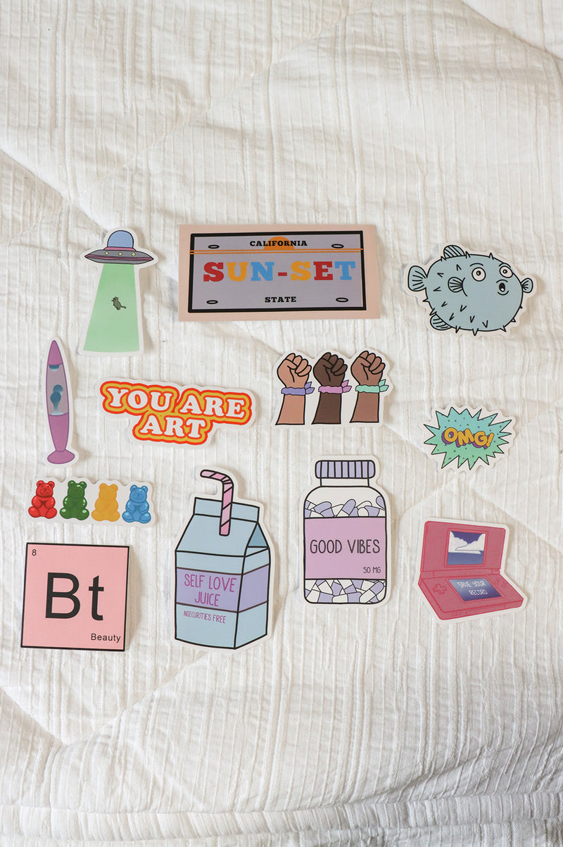 UFO sticker, "sun-set" license plate, puffer fish, lava lamp, "you are art", 3 skin tone power fist with scrunchie, "OMG" bubble, gummy bear, Bt periodic table box with "beauty" in it, "Self Love Juice" Milk Carton, "Good Vibe" vitamin bottle, DSI in pink